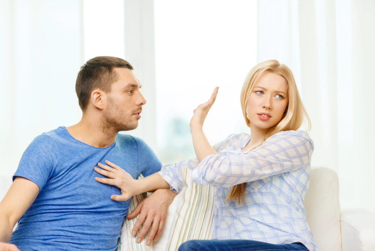 A woman sitting on a couch raises her hand and turns away from a man next to her, who is reaching toward her; her response suggests she may be reacting based on her attachment style.