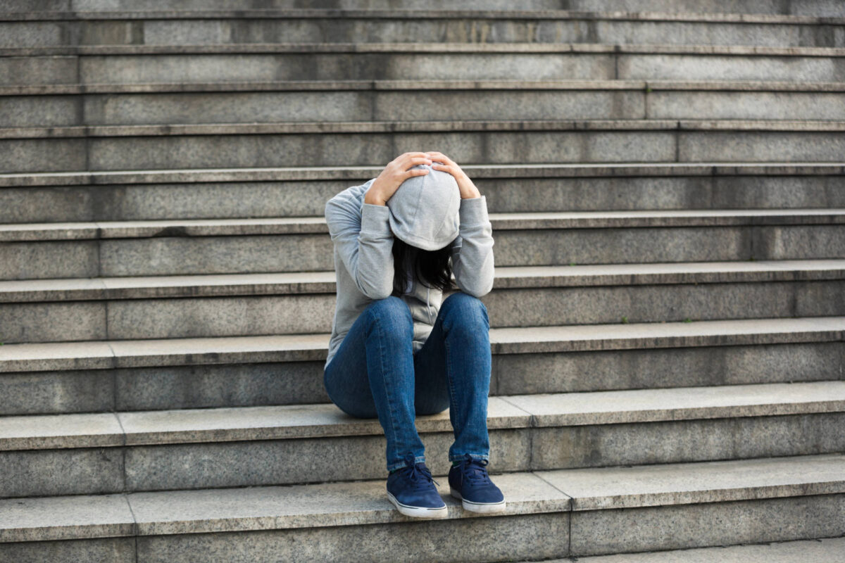 A person in a gray hoodie and blue jeans sits on outdoor stone steps with head down and hands on head, appearing distressed, as if discovering shame or wrestling with toxic shame in a moment of quiet reflection.