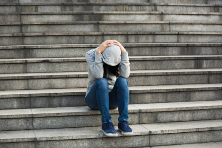 A person in a gray hoodie and blue jeans sits on outdoor stone steps with head down and hands on head, appearing distressed, as if discovering shame or wrestling with toxic shame in a moment of quiet reflection.