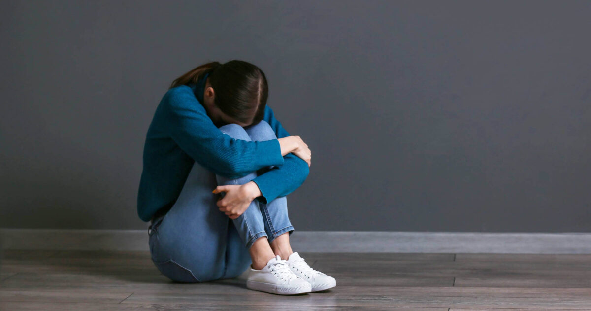 woman-hugging-legs-staying-small-1800-sm A person sits on the floor with their knees pulled to their chest and head down, arms wrapped around their legs, reflecting our tendency to shrink into ourselves and stay small instead of fully expressing who we are after trauma.