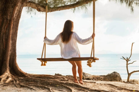A person sits on a wooden swing hanging from a tree by the ocean, using pendulation and titration for processing anger.
