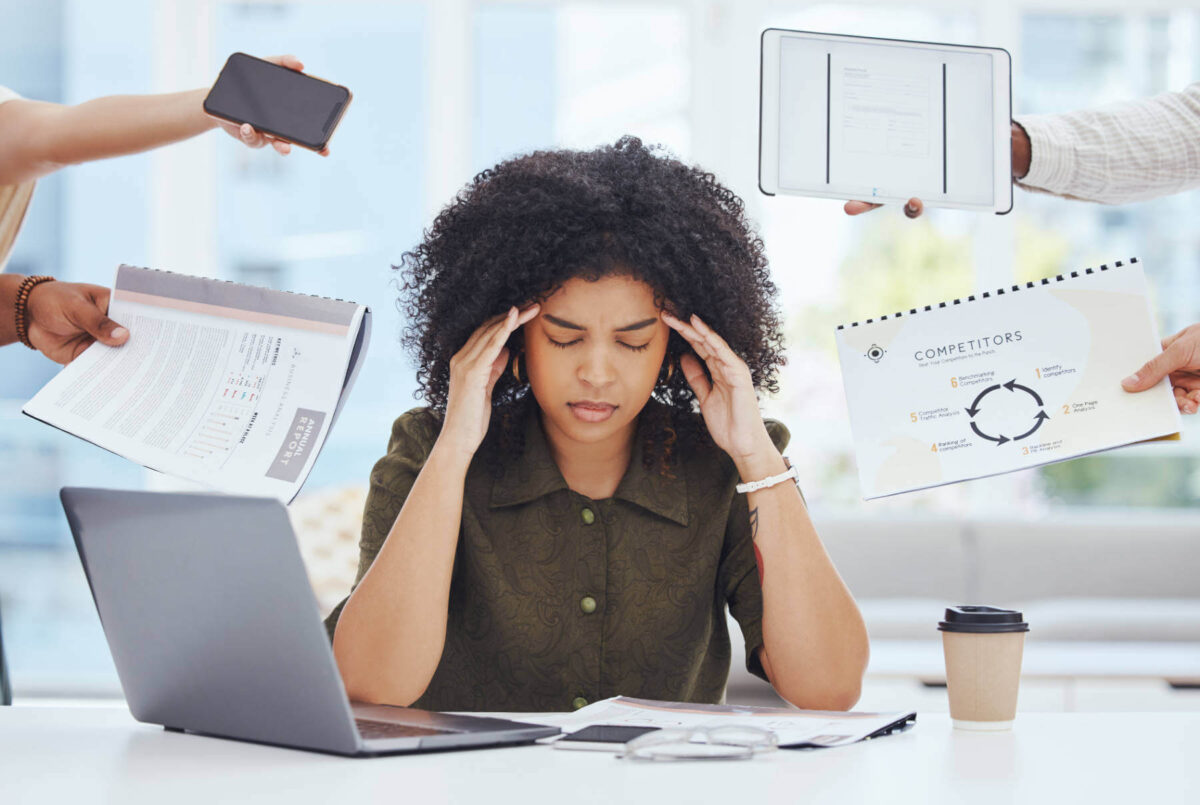 woman-stressed-burnout-overworked-1800-sm A woman sits at a desk with her hands on her temples, looking stressed, as workaholism takes its toll. Around her is a phone, tablet, and documents, encouraging constant productivity and busyness.