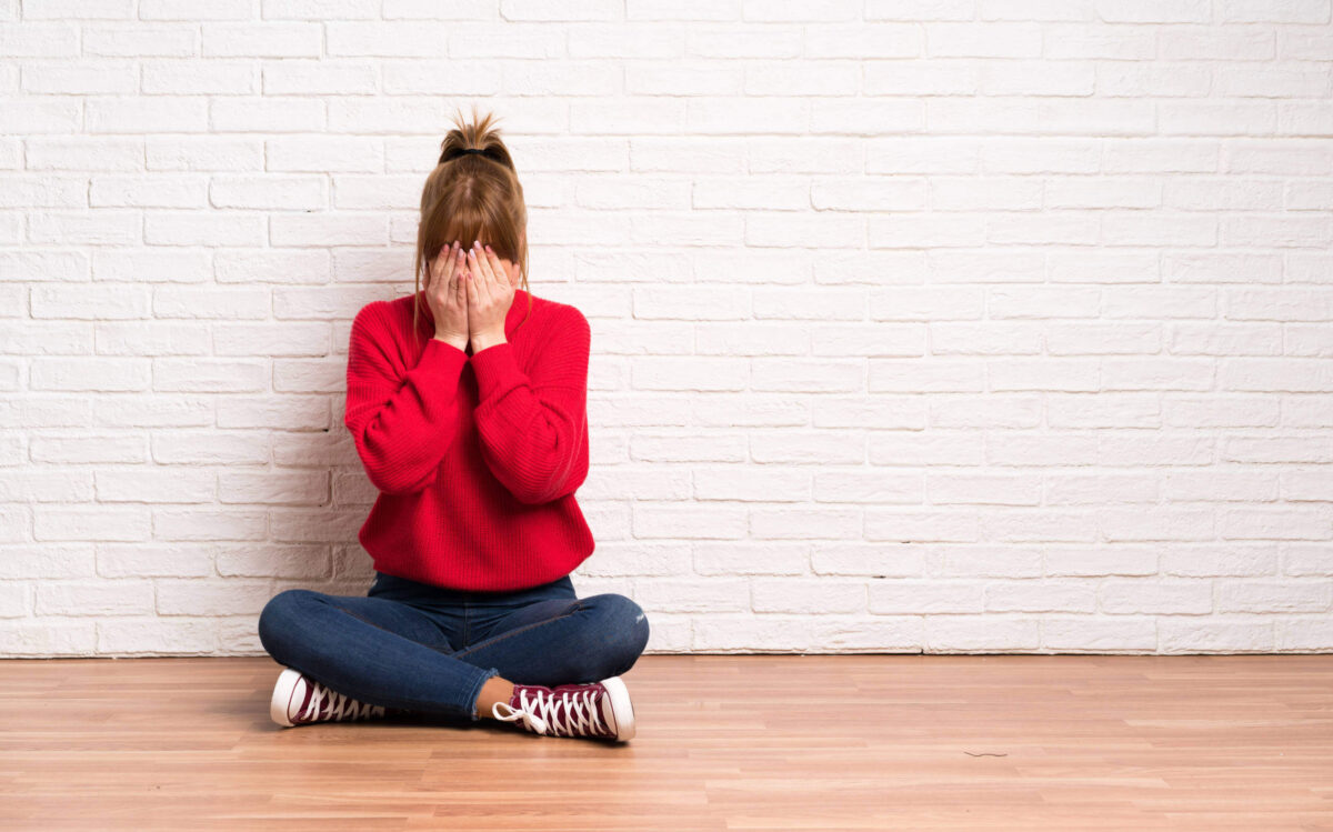 A person in a red sweater and jeans sits cross-legged on a wooden floor against a white brick wall, covering their face with both hands, showing the struggle of managing uncomfortable feelings.